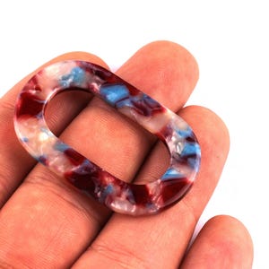Peut inclure: Un lien ovale en acrylique dans les tons de rouge, blanc et bleu. Le motif marbré est visible sur tout le lien. Le lien est tenu dans une main sur fond blanc. Ce lien décoratif peut être utilisé pour la fabrication de bijoux.