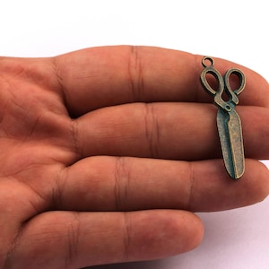 May include: A small, antique bronze-colored scissors charm. The charm has a loop at the top for attaching to a necklace or bracelet. The scissors are detailed and have a vintage aesthetic, perfect for jewelry making or crafting.