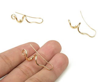 May include: Gold-colored, abstract-shaped earrings. The earrings have a curved design with a spiral at the top and a hook for the ear. One earring is held in a hand.