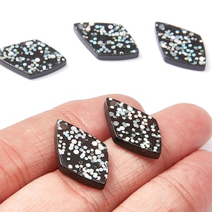 May include: Black and silver glitter rhombus-shaped pendants. The pendants have a small hole at the top for stringing. The glitter is a mix of silver and white, set against a black background. The pendants are shown on a white background.