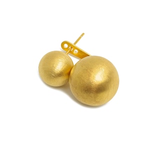 Puede incluir: Un par de pendientes dorados de doble esfera. Los pendientes tienen un poste y una barra de conexión, con una esfera más pequeña y otra más grande. Las esferas tienen un acabado mate texturizado, sobre un fondo blanco.