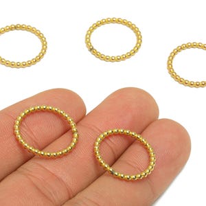 Puede incluir: Primer plano de varios anillos dorados. Los anillos están adornados con pequeñas cuentas espaciadas uniformemente, creando una apariencia texturizada y con cuentas. Dos anillos se sostienen en una mano, mientras que otros tres están dispersos sobre una superficie blanca.