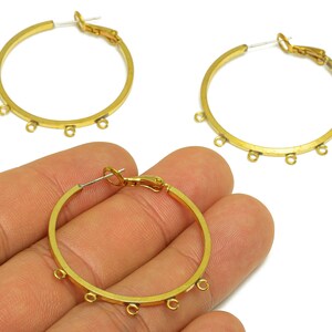 May include: Gold-toned hoop earrings with small loops for attaching charms. The earrings have a simple, circular design with a clasp closure. One earring is held in a hand, showcasing the design.