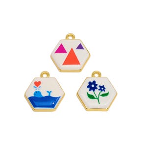 Op de afbeelding: Drie zeshoekige metalen bedels met goudkleurige randen en witte emaille centra. Eén bedel heeft een patroon van roze, oranje en paarse driehoeken. Een andere toont een blauwe walvis met een rood hart. De derde toont een blauwe bloem met bladeren.