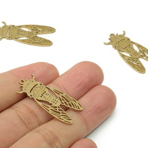 May include: Three gold-colored cicada charms. The charms are detailed with etched wings and body segments. One charm is held in a hand, while two others are scattered on a white background. These charms are suitable for jewelry making or crafting.