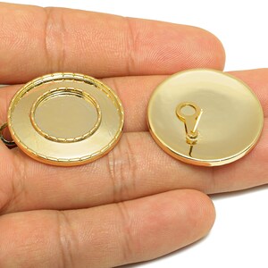 May include: Two gold-colored, round jewelry findings. One has a recessed center and a loop for attachment. The other has a loop on the back. These findings are likely used for creating jewelry such as earrings or pendants.