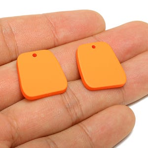 May include: Two orange rectangular pendants with rounded corners and a small hole at the top. The pendants are held in a person's hand. The pendants have a smooth, matte finish and are a vibrant shade of orange.