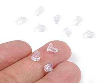 May include: A collection of clear, rubber earring backs. Several are scattered on a white surface, while three are held between fingers. The earring backs are small, with a tapered shape and a hole for the earring post.