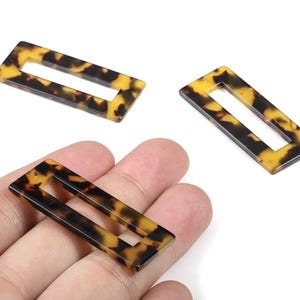 Puede incluir: Cuentas rectangulares de acrílico con estampado de carey. Las cuentas tienen forma rectangular hueca con esquinas redondeadas. Los colores son una mezcla de marrón, negro y amarillo.