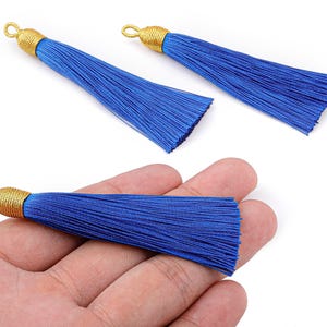 May include: Three blue tassels with gold-colored caps and loops. One tassel is held in a hand. The tassels are made of many strands of blue thread. The gold caps are tightly wound with gold-colored thread.