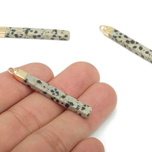 May include: Dalmatian jasper gemstone pendants with gold-colored caps and loops. The pendants are rectangular with a speckled pattern of black spots on a light gray background. The pendants are shown in a hand.