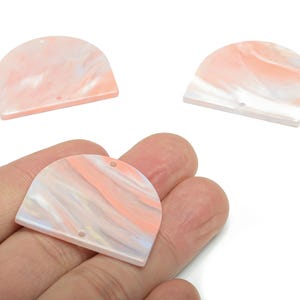 Puede incluir: Tres colgantes semicirculares de acrílico veteado en tonos melocotón, blanco y azul claro. Cada colgante tiene un pequeño agujero para ensartar. Un colgante se sostiene en una mano.