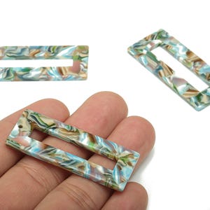 May include: Rectangular acrylic beads with a marbled pattern in shades of blue, green, brown, and cream. The beads have a rectangular shape with a cutout in the centre. The beads are shown in a close-up shot.