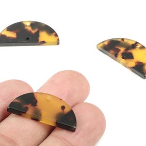 Può includere: Tre componenti di gioielleria a forma di semicerchio con motivo tartarugato. I componenti sono un mix di tonalità marrone scuro e giallo-arancio. Un componente è tenuto dalle dita di una persona. Ogni componente ha un piccolo foro per l'infilatura.