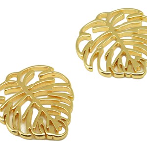 May include: Two gold-colored metal charms shaped like monstera leaves. The leaves are cut out, creating a delicate and intricate design.