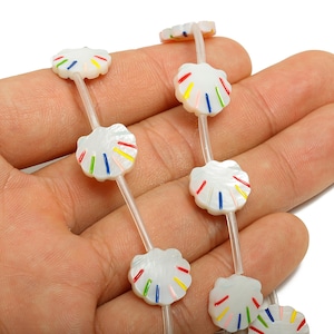 Puede incluir: Cadena de cuentas blancas en forma de concha con rayas de colores. Las cuentas están ensartadas en un cordón transparente y fino. Las cuentas se sostienen en una mano.