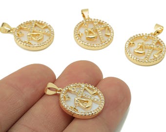 May include: Gold-colored Libra zodiac pendant with a white center and a halo of small, clear crystals. The pendant features the scales of justice symbol. The image shows multiple pendants, with one held in a hand.
