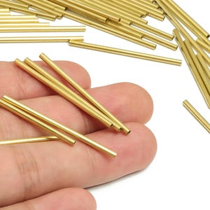 May include: A collection of gold-coloured, hollow metal tubes. Some are held in a hand, others are scattered on a white surface. The cylindrical tubes are likely components for jewellery making or other crafts. Each tube is approximately 2.5 cm long.