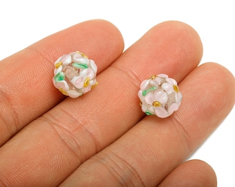 May include: Two round, floral-shaped beads. The beads are clear with pink, white, green, and yellow accents. The beads are held in a person's hand against a white background. These beads are suitable for jewelry making.