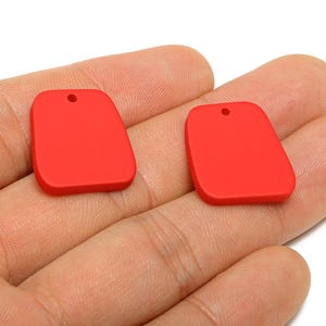 Peut inclure: Deux pendentifs rouges rectangulaires aux coins arrondis et un petit trou en haut. Les pendentifs sont tenus dans une main sur fond blanc. Ces pendentifs sont adaptés à la fabrication de bijoux.