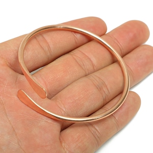 Puede incluir: Un brazalete de puño abierto de color cobre. El brazalete tiene un diseño simple y minimalista con extremos redondeados. El diseño abierto permite un fácil ajuste y uso. El brazalete se muestra en una mano.