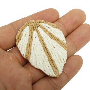 May include: A leaf-shaped decorative item with a woven texture. The leaf features alternating sections of beige and white, creating a striped pattern. The item is held in a hand against a white background.