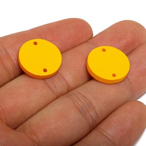 以下が含まれることがあります： 明るい黄色の円形クラフトパーツが2つ、それぞれに小さな穴が2つあり、手に持たれています。これらの平らで丸いパーツは、ジュエリー作りやその他のDIYプロジェクトに使用される可能性があります。色は鮮やかで、単色の黄色です。