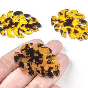 May include: Three leaf-shaped decorative pieces with a tortoiseshell pattern. The leaves are a mix of yellow and dark brown, with a glossy finish. One leaf is held in a hand, while the other two are on a white surface. These are likely jewelry components.