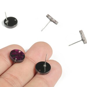 May include: A collection of round stud earrings. Some are black with a dark pink floral design, while others are black. The earrings have silver posts. The earrings are displayed on a white background.