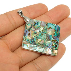 May include: Square pendant with a mosaic design. The pendant features a silver bail and a surface composed of small, iridescent abalone shell pieces in shades of blue, green, and white. The pendant is held in a hand.