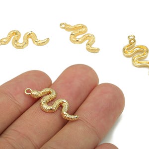 Puede incluir: Amuletos de serpiente dorados con superficie texturizada. Los amuletos tienen forma de serpiente, con un lazo en la parte superior para sujetarlos a las joyas. Varios amuletos son visibles.