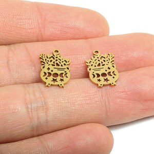 May include: Two gold-coloured cauldron charms with a witchy theme. Each charm features a cauldron shape with cut-out stars, moons, and foliage. The charms have a small loop at the top for attaching to jewellery or other crafts.