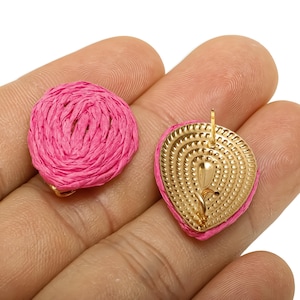 May include: A pair of pink raffia earrings. One earring is round, the other is heart-shaped with a gold-coloured metal backing. The earrings are held in a hand against a white background. The raffia is a bright, vibrant pink.