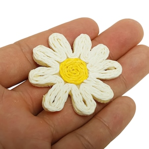 May include: A daisy-shaped craft item with white petals and a yellow centre. The flower is made of woven material and is held in a hand. This item could be used for various craft projects.