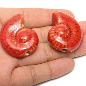 May include: Two red ceramic spiral-shaped beads with a glossy finish. The beads have a speckled pattern and a hole for stringing. The beads are held in a hand against a white background. These beads are suitable for jewelry making.