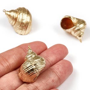 May include: Gold-colored seashell charms with textured surfaces. One charm is held in a hand, while two others are positioned nearby. Each charm has a small loop for attaching to jewelry or other crafts.