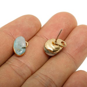 Puede incluir: Un par de pendientes de botón dorados. Cada pendiente presenta una piedra azul claro y blanca con bordes dorados. Uno es de forma ovalada, y el otro es de forma redondeada con un poste y cierre.