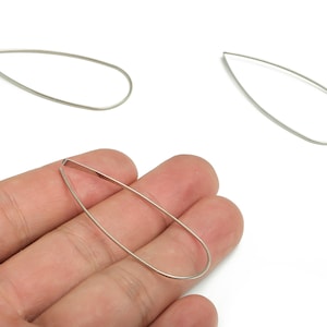 Op de afbeelding: Zilverkleurige druppelvormige oorbellen. De oorbellen zijn dun en hebben een gladde, gepolijste afwerking. Eén oorbel wordt in een hand gehouden, terwijl twee andere afzonderlijk worden weergegeven. De oorbellen zijn licht en minimalistisch.