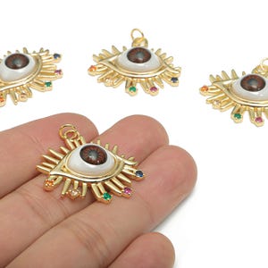 May include: Gold-colored eye-shaped pendants with a white iris and brown pupil. The pendants have colorful gemstone accents and a loop for attaching to a chain. One pendant is held in a hand.