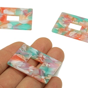 May include: Rectangular resin beads with a marbled pattern in shades of orange, teal, and pink. Each bead has a rectangular cutout in the center. The beads are shown in a hand.