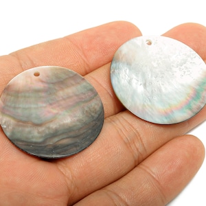 May include: Two round shell pendants with a hole at the top. One has a pearlescent white surface with green and blue hues. The other has a swirling pattern of grey, pink, and brown. Ideal for jewellery making.