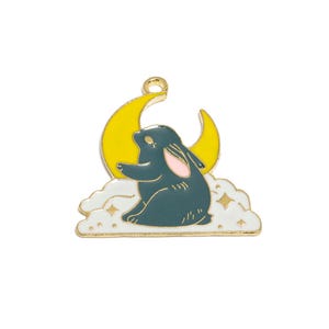 Puede incluir: Un dije dorado con un conejo verde oscuro sentado en una nube blanca con estrellas doradas. Detrás del conejo hay una luna creciente amarilla. El conejo tiene orejas internas rosas y mira hacia la izquierda.