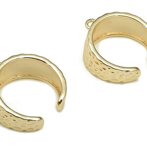 Op de afbeelding: Twee gouden metalen oormanchetten met een gehamerde textuur. Elke manchet heeft een klein lusje aan de bovenkant om aan een ketting of oorring te bevestigen.