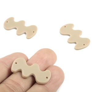 Könnte beinhalten: Drei beige, wellenförmige Bastelperlen mit jeweils zwei Löchern. Eine Perle wird in einer Hand gehalten, die anderen beiden liegen auf weißem Hintergrund. Die Perlen haben eine glatte, matte Oberfläche und bestehen wahrscheinlich aus Harz oder Ähnlichem.