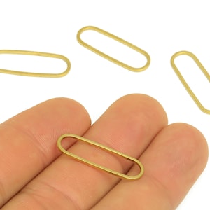 May include: Several small, gold-colored oval-shaped metal jewelry findings are displayed. One is held in a hand, while the others are scattered on a white surface. These findings are likely used for jewelry making or crafting.