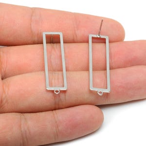 May include: Two silver-colored rectangular earring components. Each component has a rectangular frame with a small loop at the bottom and a post for piercing the ear. The earrings are shown against a light background.