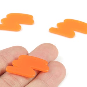 May include: Three orange abstract-shaped acrylic charms. One charm is held in a hand, while two others are scattered on a white background. The charms have a smooth, glossy finish and a small hole for stringing.