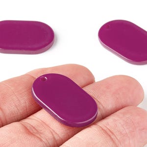 Pode incluir: Três pingentes ovais cor de ameixa, cada um com um pequeno orifício para enfiar. Um é segurado na mão, os outros em fundo branco. Estes pingentes são adequados para fazer joias, medindo cerca de 2,5 cm de comprimento.