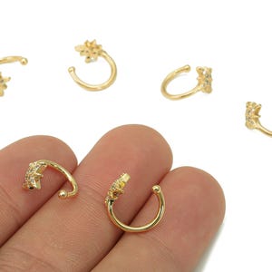 Puede incluir: Puños de oreja dorados con diseño de estrella. Los puños de oreja son pequeños, curvados y presentan un grupo de pequeñas piedras preciosas transparentes. Las joyas se muestran sobre un fondo blanco.