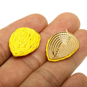 May include: A pair of yellow raffia earrings. One earring is a solid, woven design, while the other reveals a gold-coloured metal backing with a post for piercing. The earrings are held in a hand.